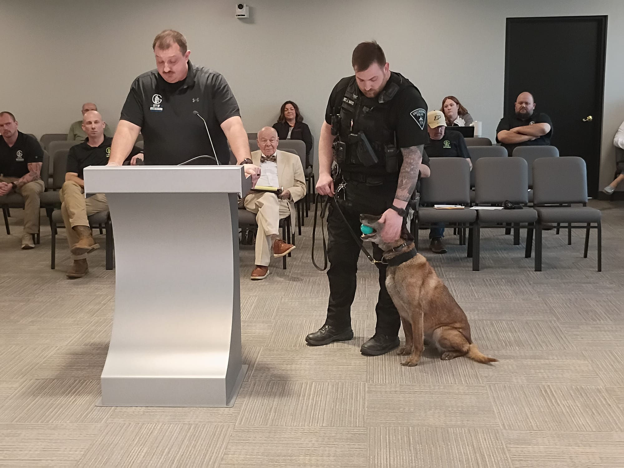 Two GPD Officers in black uniforms, one speaking at a podium, the other holding K-9 officer Kato's leash. Kato is a brown Belgian Malinois chewing a turquoise-colored ball.