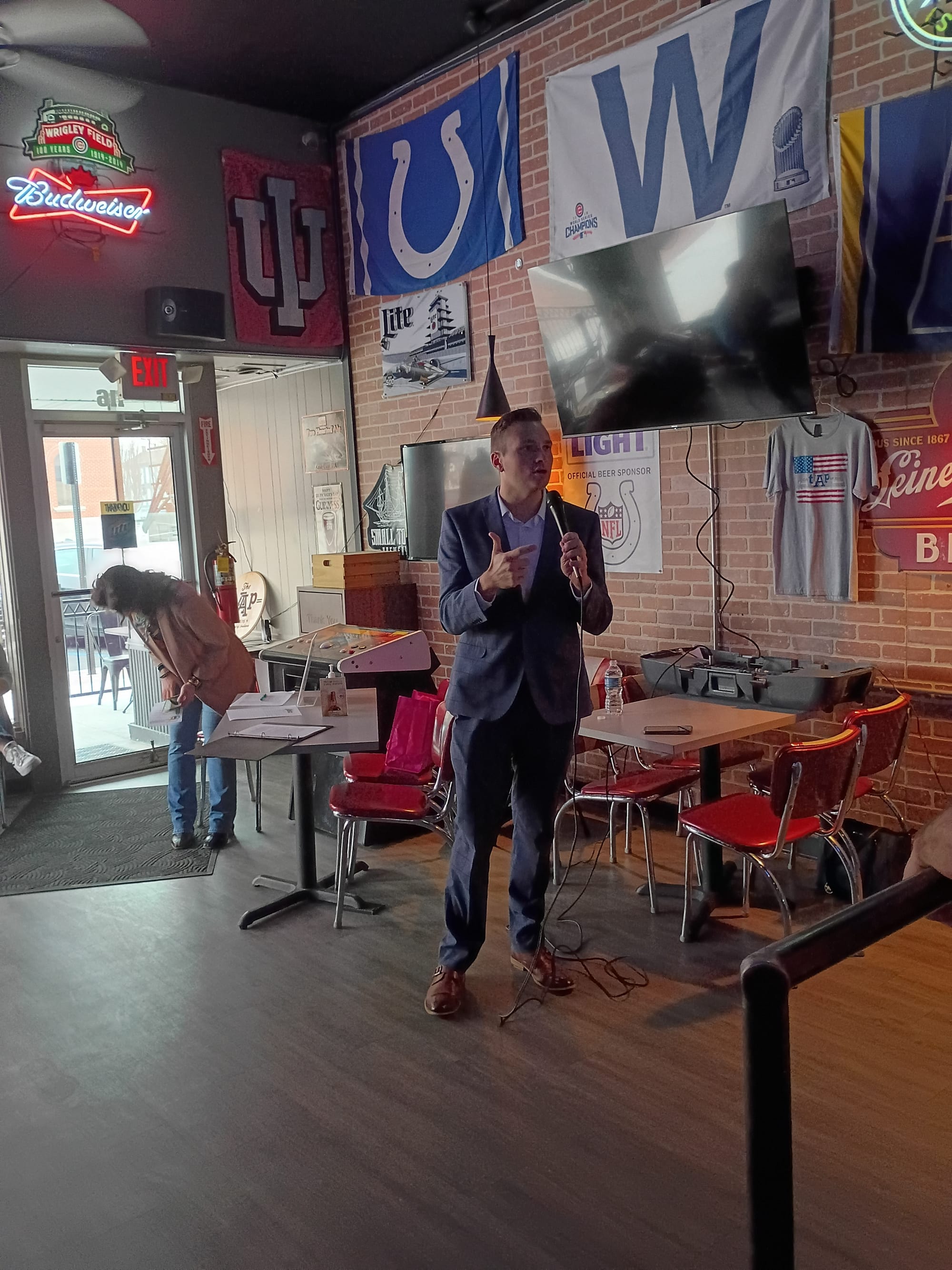 Greensburg Mayor Joshua Marsh standing inside The Taap giving the 2025 State of the City address during the Chamber of Commerce Leaders and Lagers event. There is sports memorabilia on the brick wall behind Mayor Marsh. Photo by Noelle Maxwell.