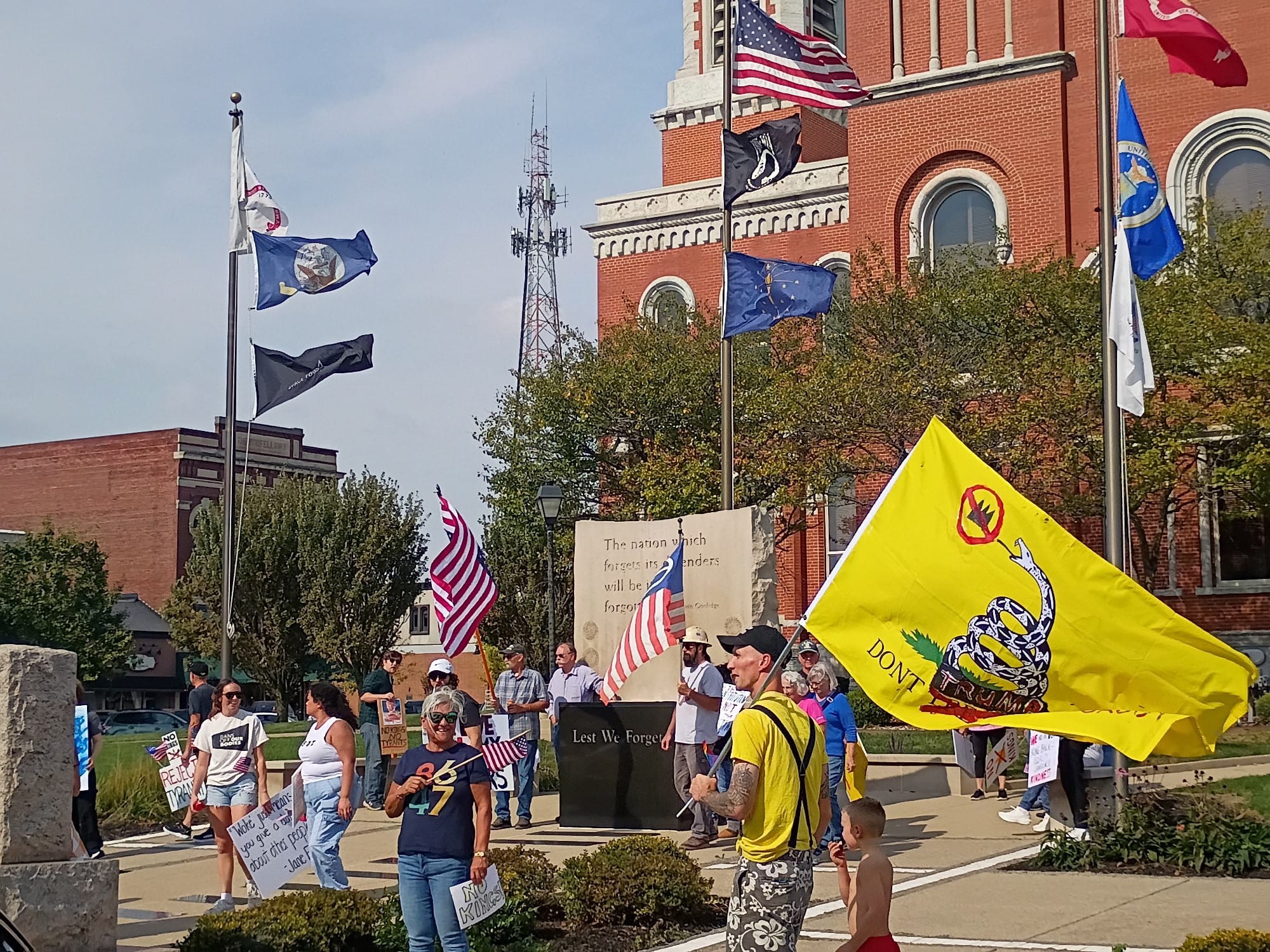 Peaceful protestors rallying at the October 2025 No Kings Day protest in Greensburg. One protestor is carrying a variant of the Gadsden Flag depicting a rattlesnake facing a "No Kings" symbol - a crown with a universal "no" symbol over it - and the word "Tread" has been crossed out and replaced with "Trump." Several protestors are carrying signs or American flags.