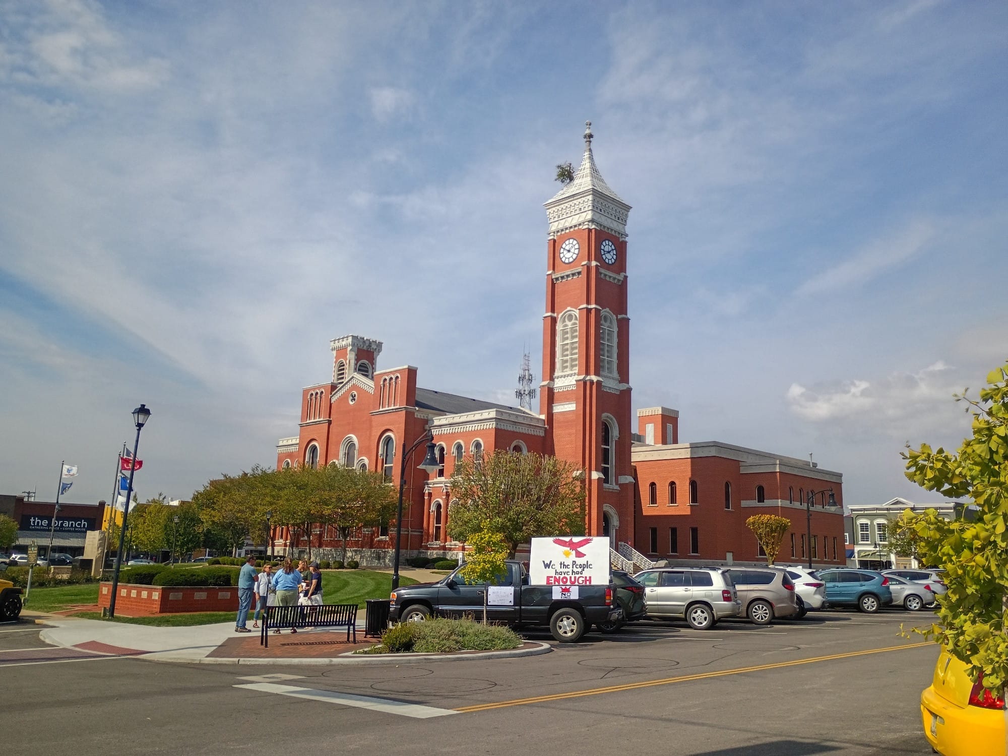A photo of the Decatur County Courthouse in Greensburg, Indiana. The courthouse is a red brick building with a white roof and trim. There's a clock tower with a tree growing out of it. There are several cars parked in front of the building and a small group of people standing on the sidewalk. One of the parked cars is a black pick-up truck with a large sign in the back depicting a drawing of a Cardinal, Indiana's state bird. Below the cardinal, the sign reads "We the People have had ENOUGH." Photo by Noelle Maxwell.