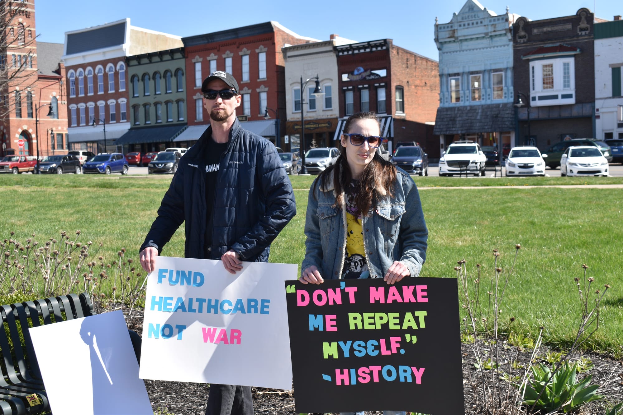 Two protesters holding signs. One protester is a man with a goatee wearing a baseball cap, sunglasses and dark colored jacket. His sign says "Fund healthcare not war" in blue and hot pink letters on a white background. One protester is a woman wearing sunglasses, a denim jacket and a Pikachu t-shirt. Her sign says "Don't make me repeat myself. -History," in hot pink, blue and lime green letters on a black background. Photo by Noelle Maxwell.