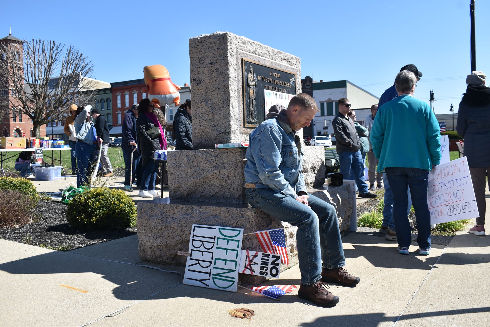 A man participating in the protest sitting, with his sign next to him, at the start of the event. He's wearing a denim jacket with jeans and brown work/hiking boots. His sign has two United States flags attached to it and reads "No Kings, Defend Liberty." Photo by Noelle Maxwell.