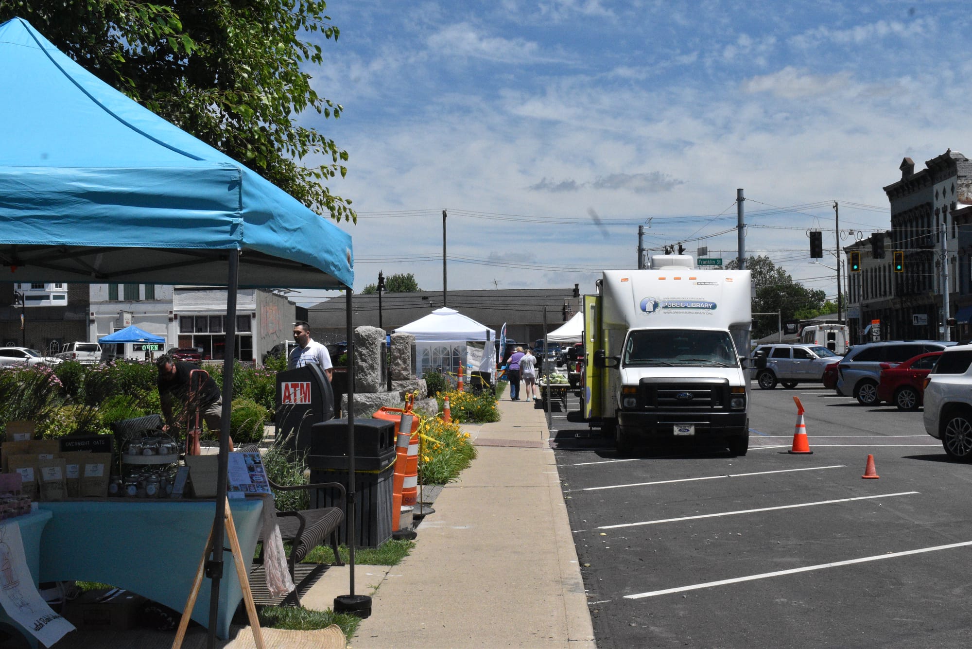 Greensburg, IN farmers' market, June 2026. The sky is blue with a few clouds, there is a blue tent selling baked goods at the front of the photo, a white bus-like vehicle that serves as the library's book mobile further back, and white tents selling other goods in the background. Photo by Noelle Maxwell.
