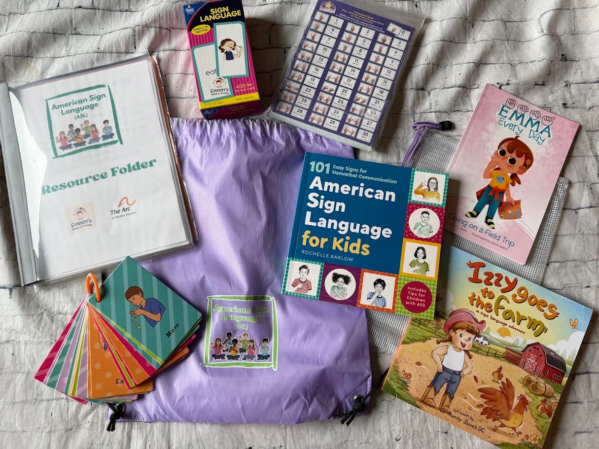 An American Sign Language kit, one of several types of sensory kits available at the Greensburg-Decatur County Public Library. The kit contains an ASL resource folder, at least two sets of ASL flash cards, a copy of "101 Easy Signs for Nonverbal Communication: American Sign Language for Kids," by Rochelle Barlow, a copy of "Emma Every Day, Going on a Field Trip," a copy of "Izzy Goes to the Farm, a Sign Language Adventure," by Dr. Mandy Jairell DC. The kit comes in a lavender drawstring bag with an image of a green square outline. Inside the green square outline is a depiction of a multi-racial group of children and the words "American Sign Language (ASL)" in green text. Photo provided.