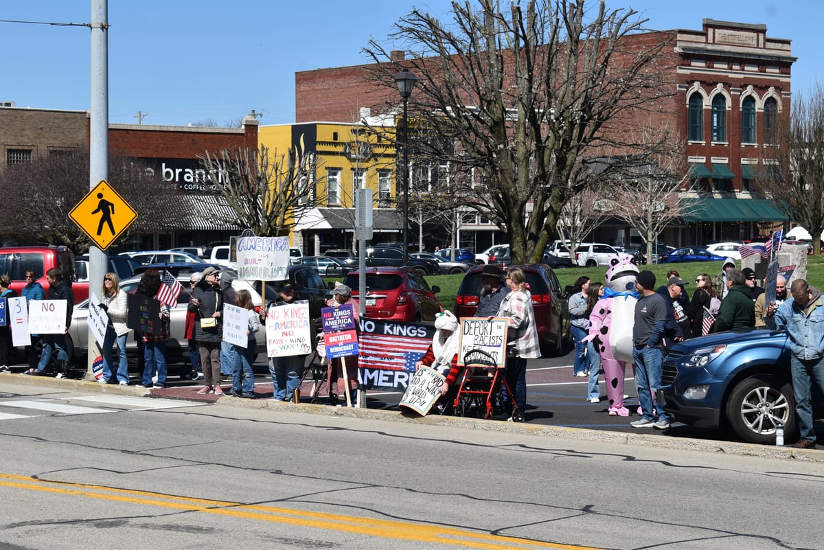 People participating in the March 28, 2026 No Kings protest in Greensburg, IN. Several people are holding signs. there is one person wearing a pink frog costume. Photo by Noelle Maxwell.