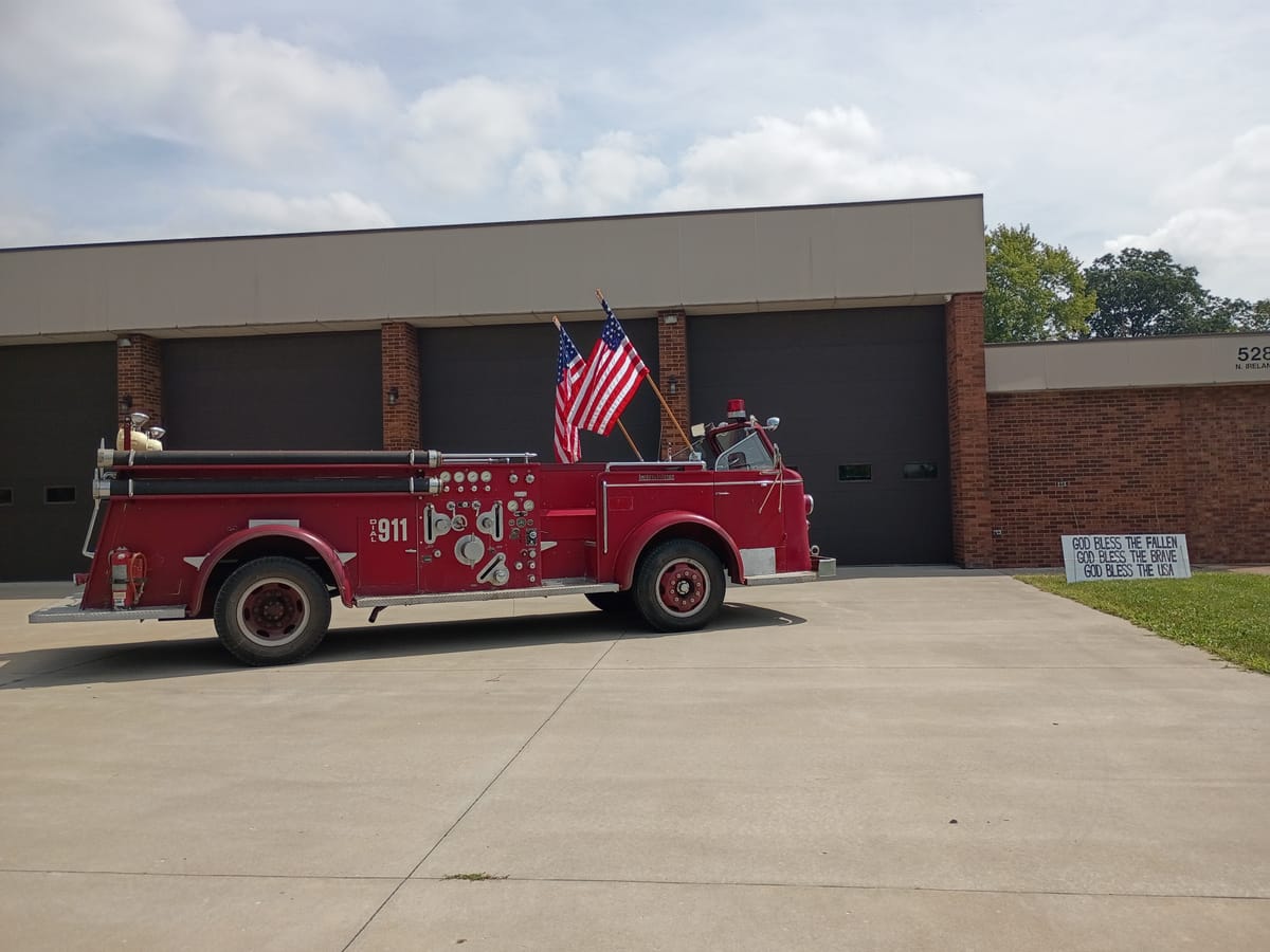 A red antique fire truck with two U.S. flags. Nearby is a sign reading "God bless the fallen. God bless the brave. God bless the USA." Photo by Noelle Maxwell.
