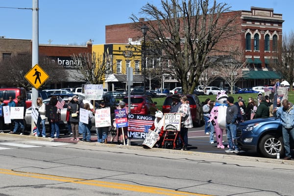 People participating in the March 28, 2026 No Kings protest in Greensburg, IN. Several people are holding signs. there is one person wearing a pink frog costume. Photo by Noelle Maxwell.